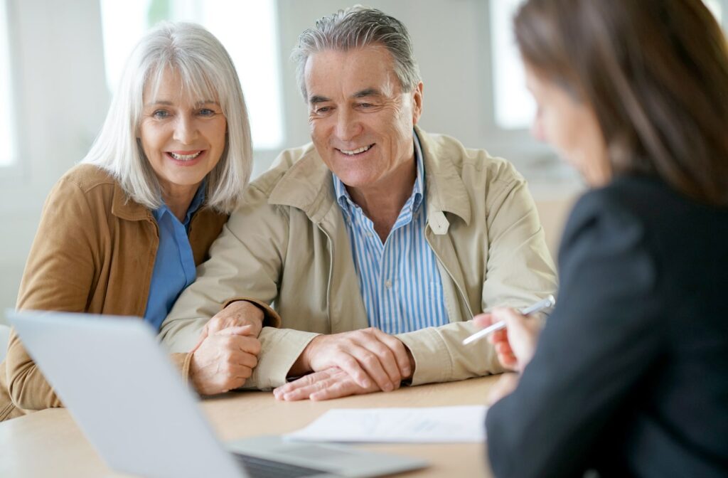 A mature smiling couple reviews information on a laptop while a female senior living community representative reviews paperwork with a pen