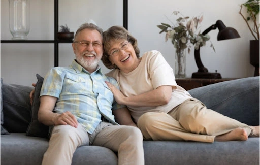 A mature couple sits on a nice grey couch as they smile and embrace each other.