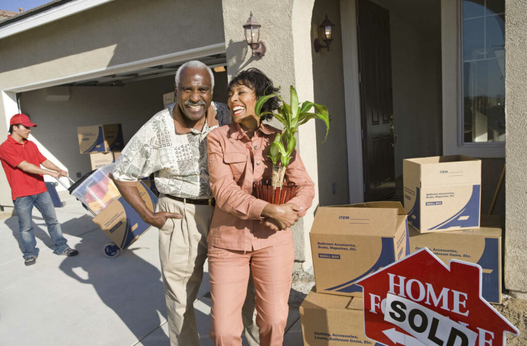 A senior couple standing and laughing next to a Home Sold sign.