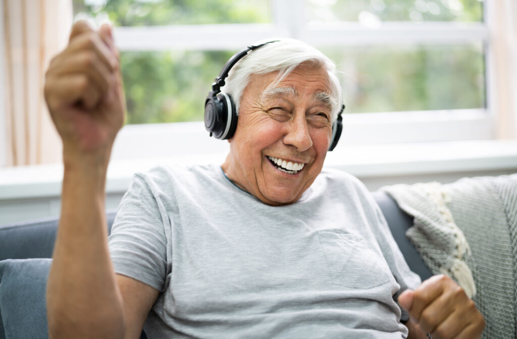 A senior man smiling and sitting on a couch wearing headphones and listening to music.