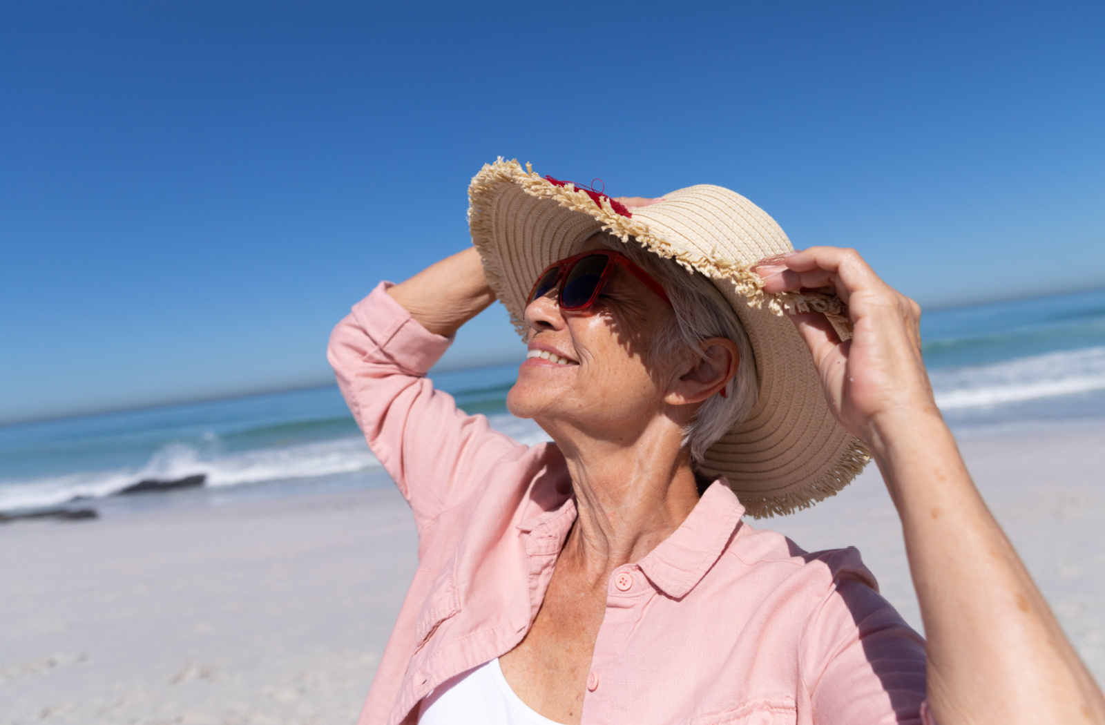 A senior woman enjoying the beach while wearing a hat and sunglasses, smiling with sea in the background.
