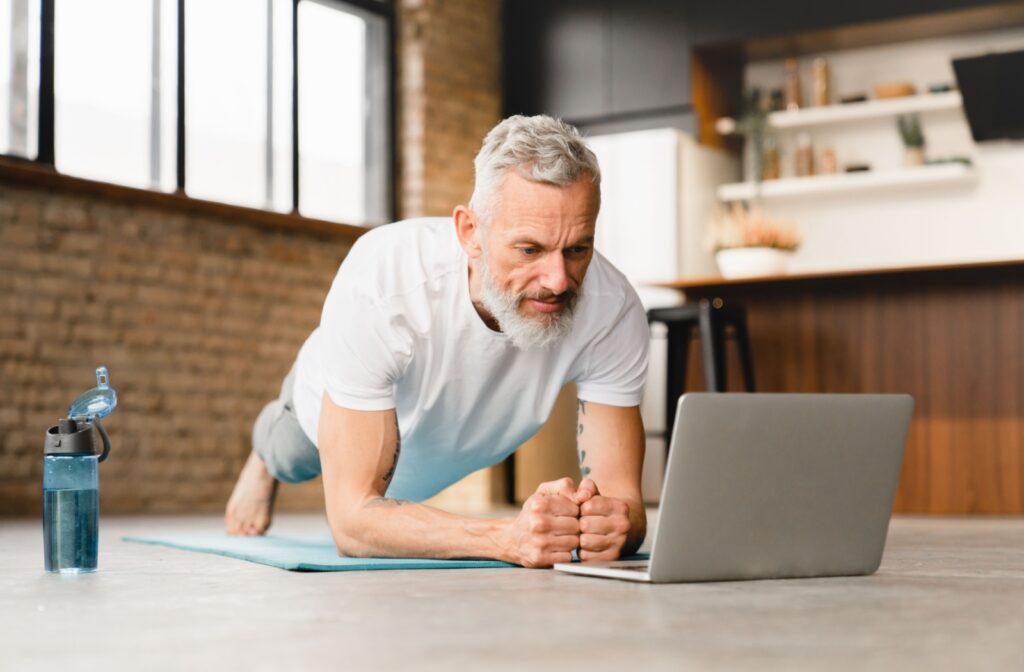 An older adult man planking on an exercise mat while looking at his laptop.