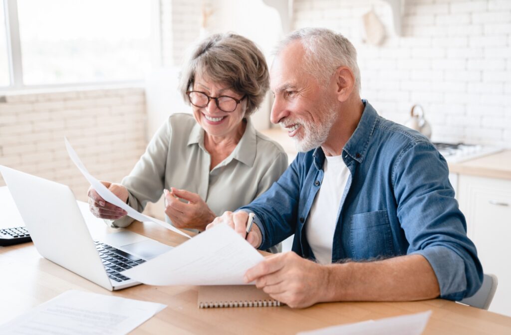 A senior couple in their home smiling with paperwork while using their laptop to research independent living communities near them.