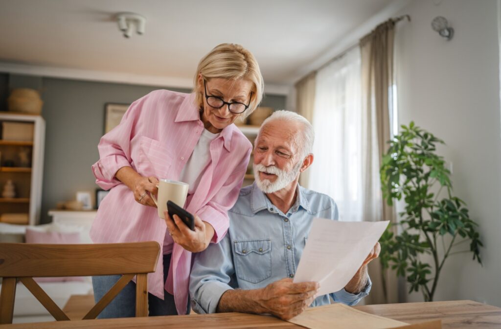 An older couple reviewing paperwork while discussing something on a smartphone in their home.