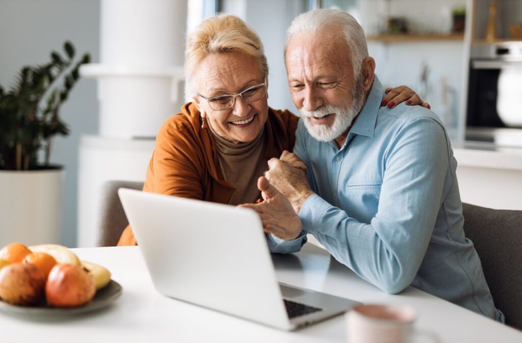 An older couple smiling and using a laptop together at a kitchen table.