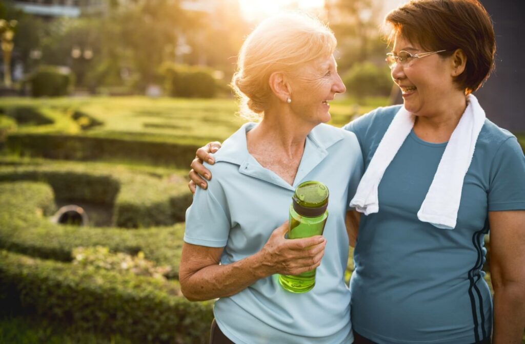 Two older adults smile in a sunny garden after exercising while the one on the left holds a water bottle.