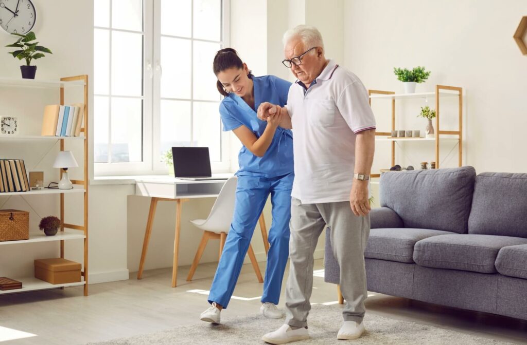 A caregiver helps an older adult safely walk across their living room in assisted living