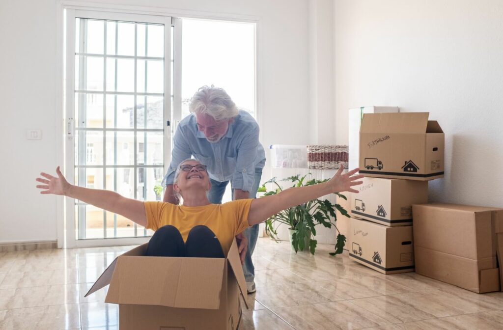 An older adult laughs and sits in a cardboard box while their spouse pushes them around a room in their new, well-lit home in senior living
