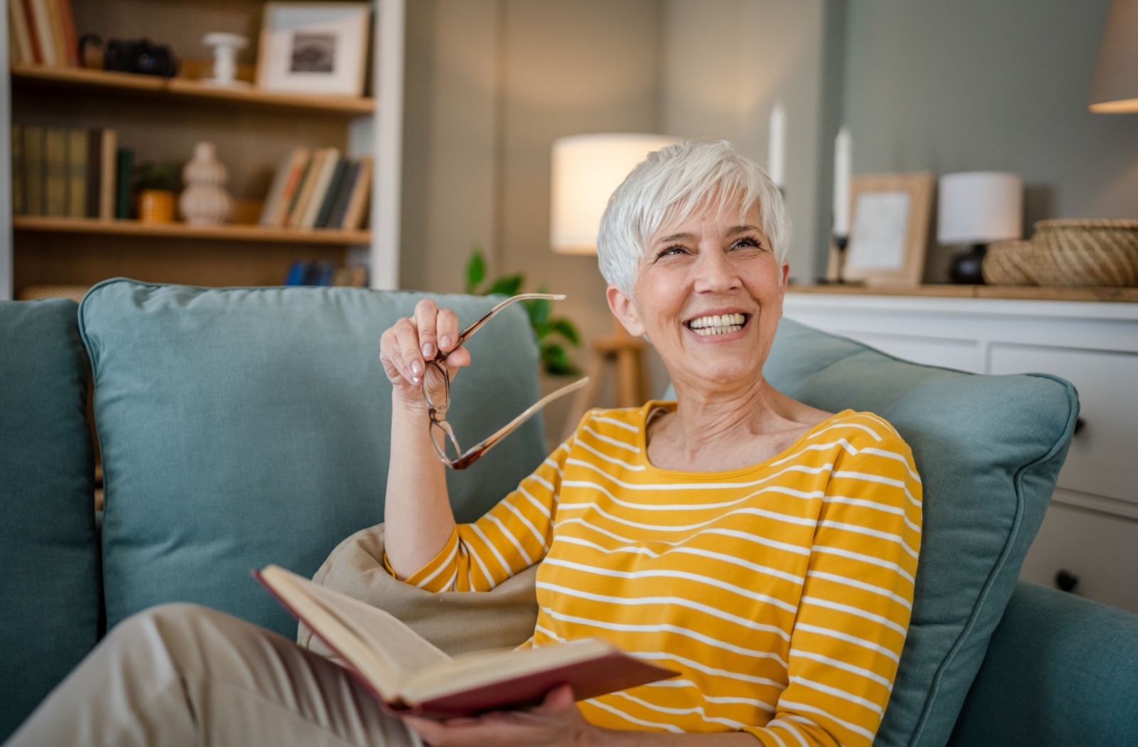 A senior smiles while reading a book and sitting on a sofa in their independent living apartment.
