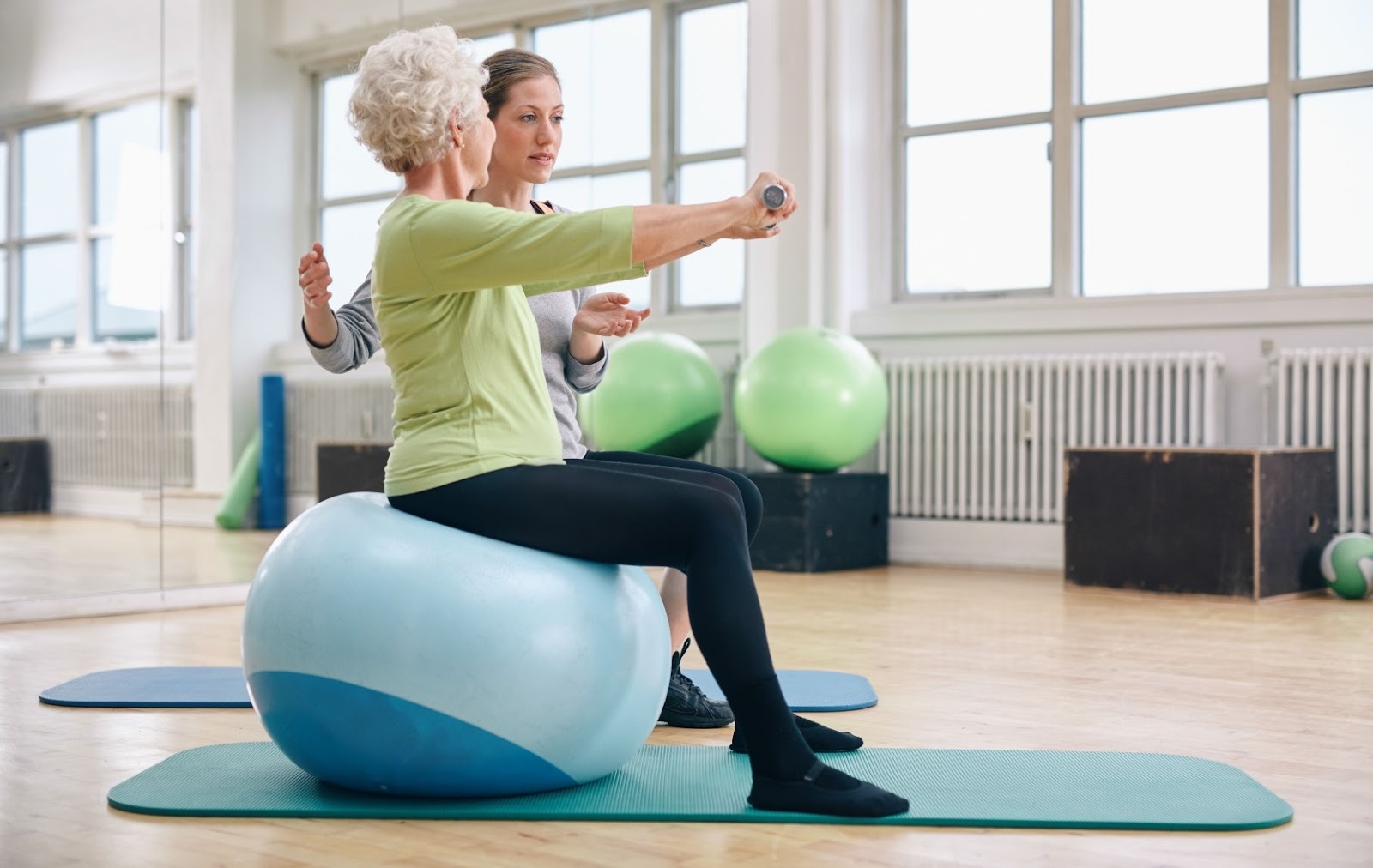 A senior uses an exercise ball with light weights to improve mobility, under the guidance of a caregiver.
